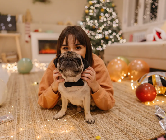 A woman lays with her pug on the floor, enjoying the cozy vibe of Christmas decorations as they welcome the New Year.