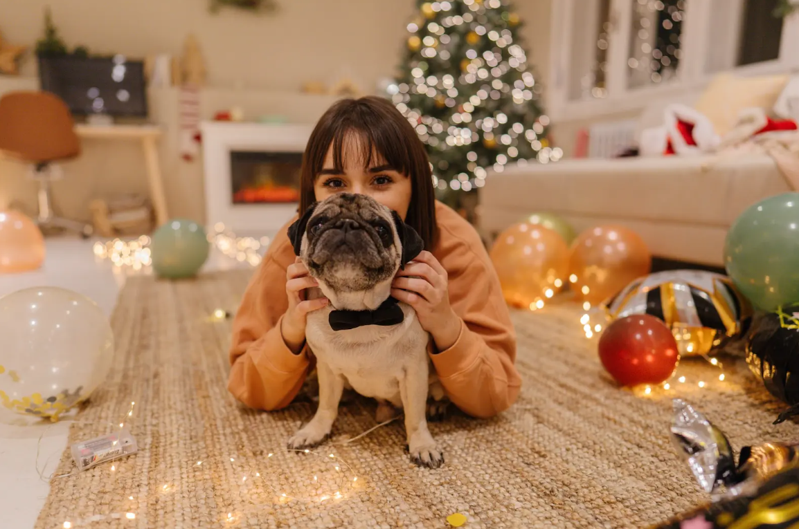 A woman lays with her pug on the floor, enjoying the cozy vibe of Christmas decorations as they welcome the New Year.
